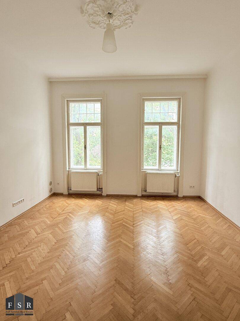 Geräumiges Zimmer mit Fischgrätparkett, hohen Decken und Stuckverzierung an der Decke.