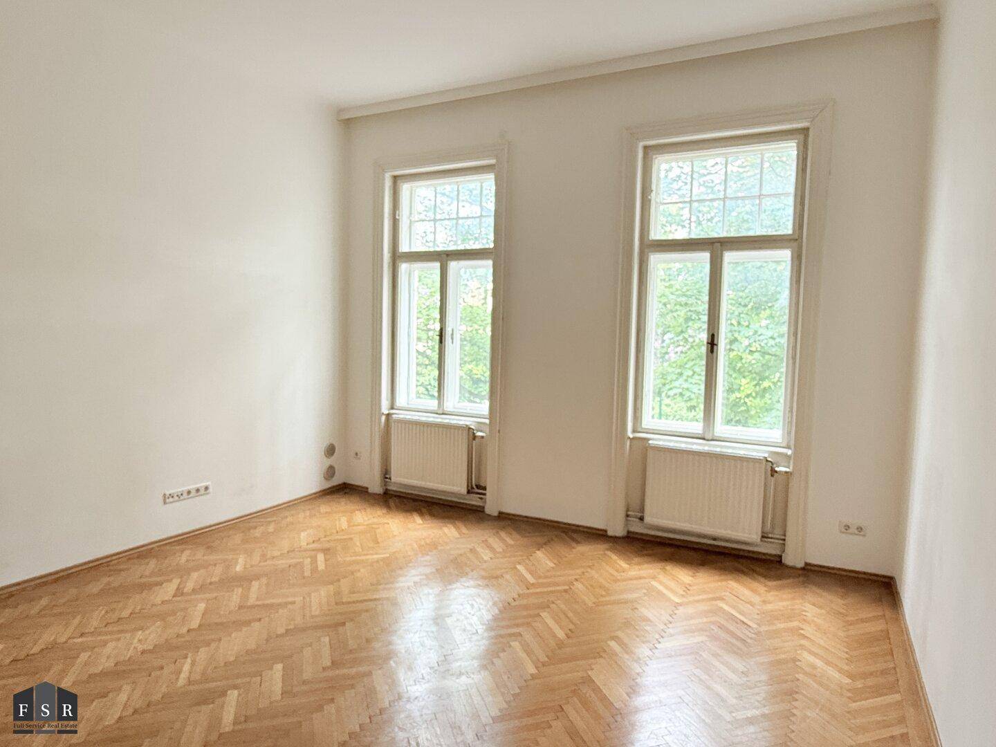 Helles Zimmer mit Fischgrätparkett, hohen Decken und zwei großen Fenstern.