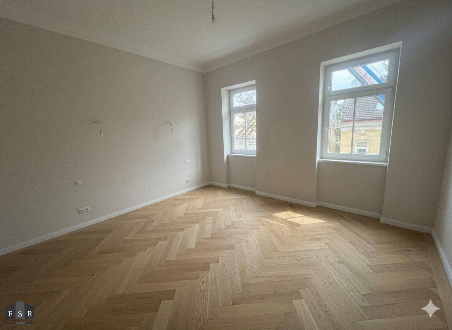 Heller Raum mit Fischgrätparkett und zwei Fenstern, die viel Sonnenlicht in den Raum lassen.