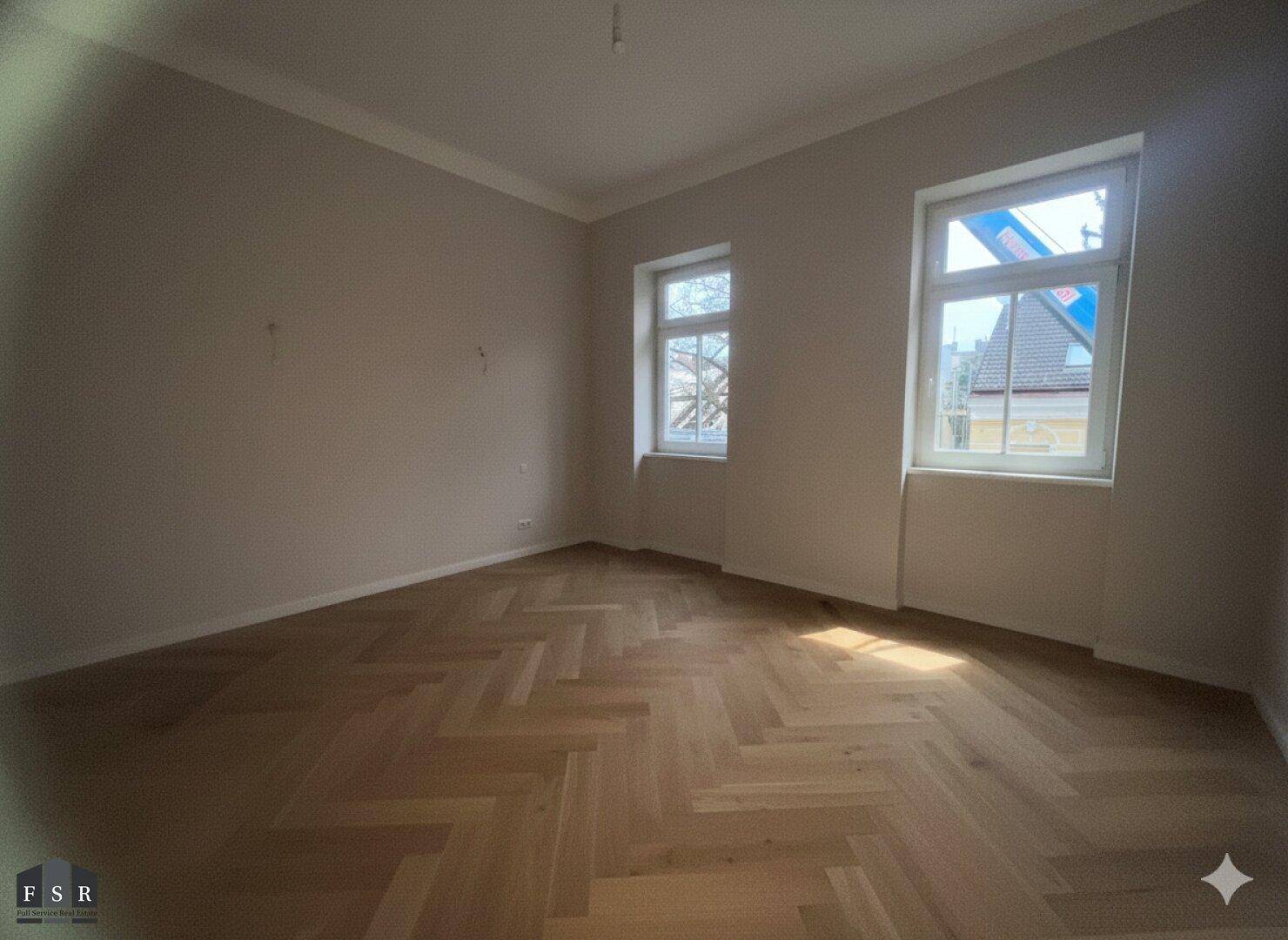 Heller Raum mit Fischgrätparkett und zwei Fenstern, die viel Sonnenlicht in den Raum lassen.