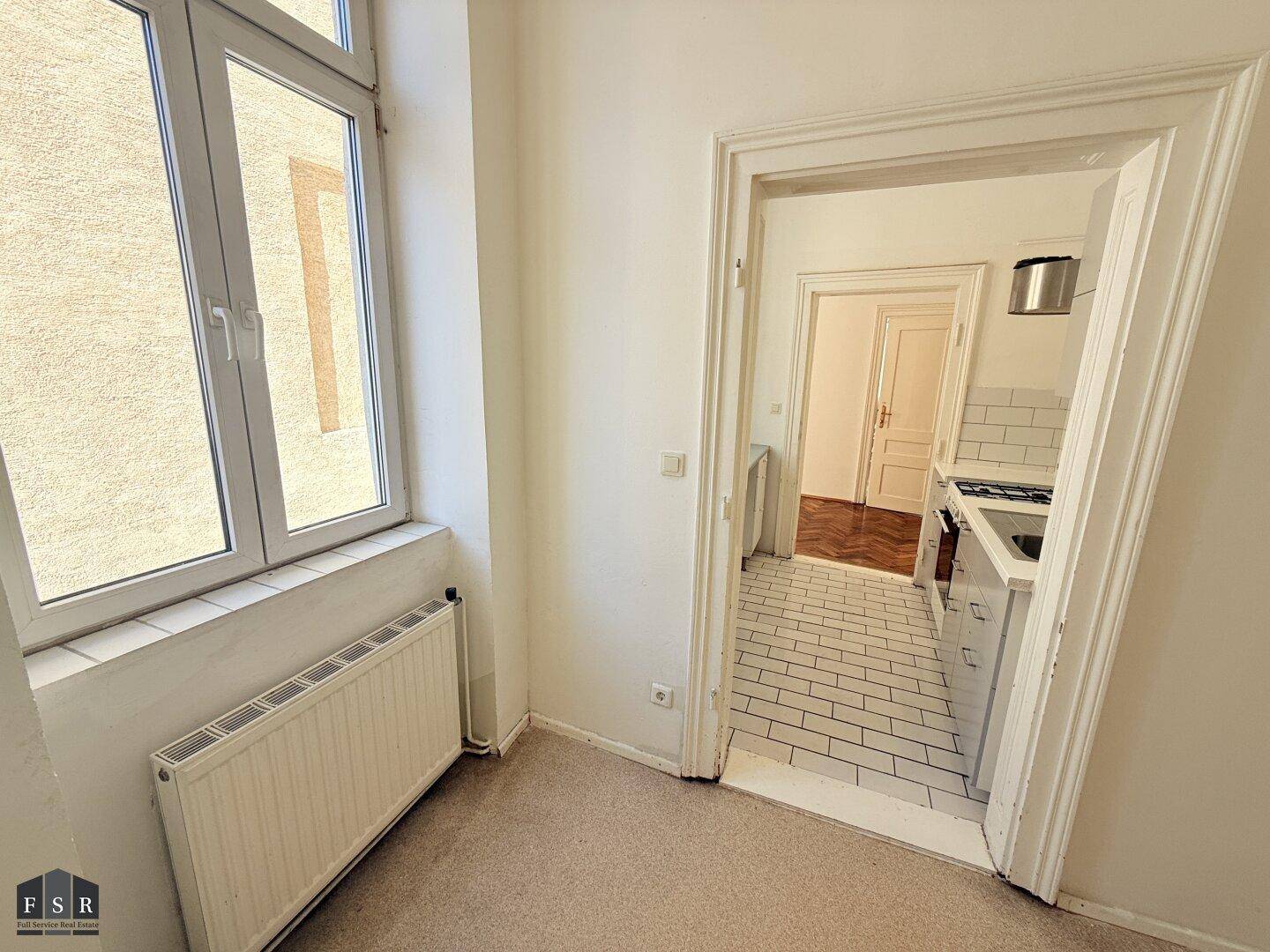 Raum mit Teppichboden und Zugang zu einer gefliesten Küche, beleuchtet durch ein Fenster und in gutem Zustand.