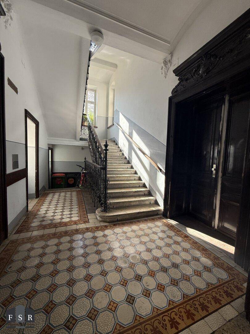 Historisches Treppenhaus mit kunstvollem Geländer, gemusterten Fliesen und viel Tageslicht, das den Altbaucharakter betont.