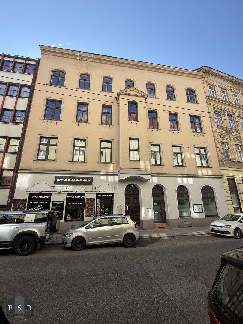 Klassische Fassade eines Mehrfamilienhauses mit Geschäften im Erdgeschoss und parkenden Autos auf der Straße.