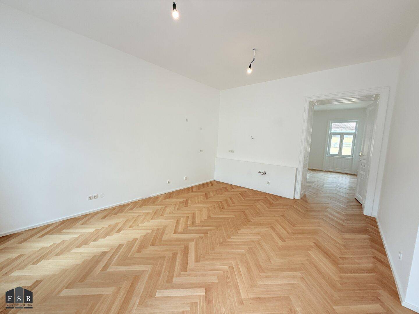 Helles Zimmer mit Fischgrätparkett, hohen Decken und einem offenen Durchgang zu einem weiteren Raum.