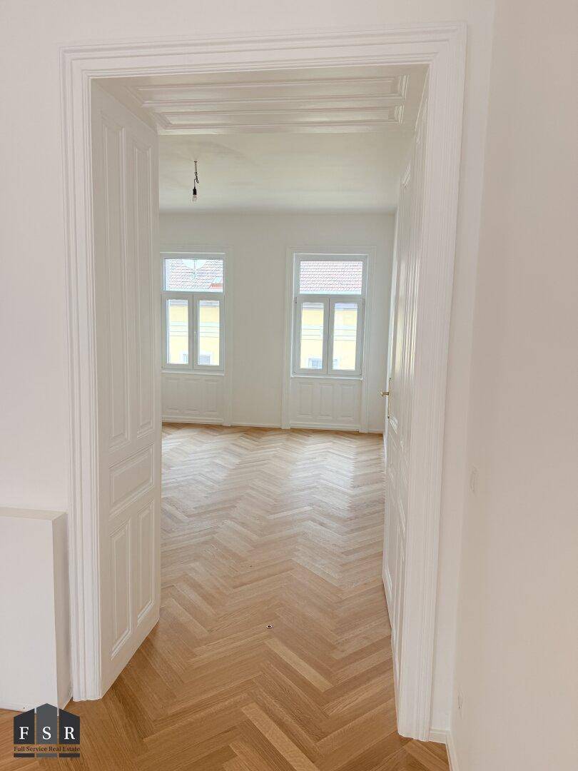 Blick durch eine klassische Tür in ein helles Zimmer mit Fischgrätparkett und Stuckdecke.