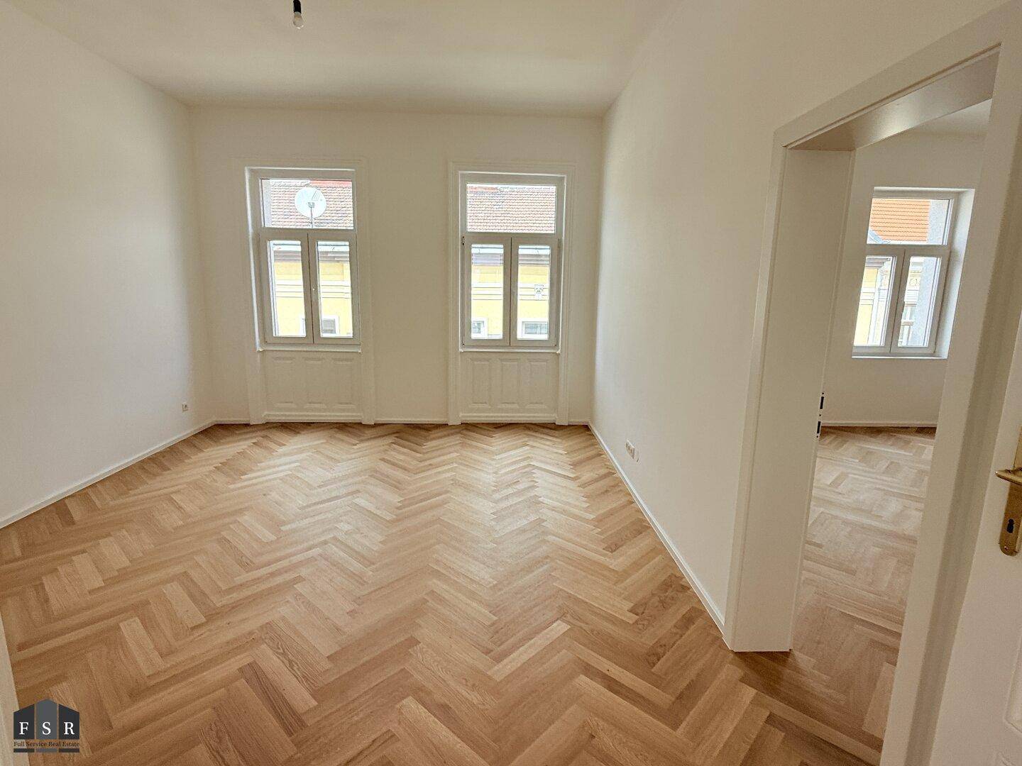 Geräumiges Zimmer mit Fischgrätparkett, hohen Decken und einem offenen Durchgang zu einem weiteren Raum.
