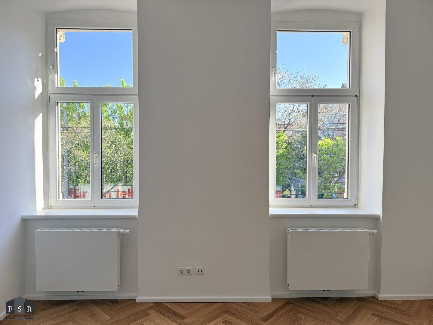 Zwei große Fenster mit Blick auf grüne Bäume und Heizkörper darunter in einem hellen Raum.