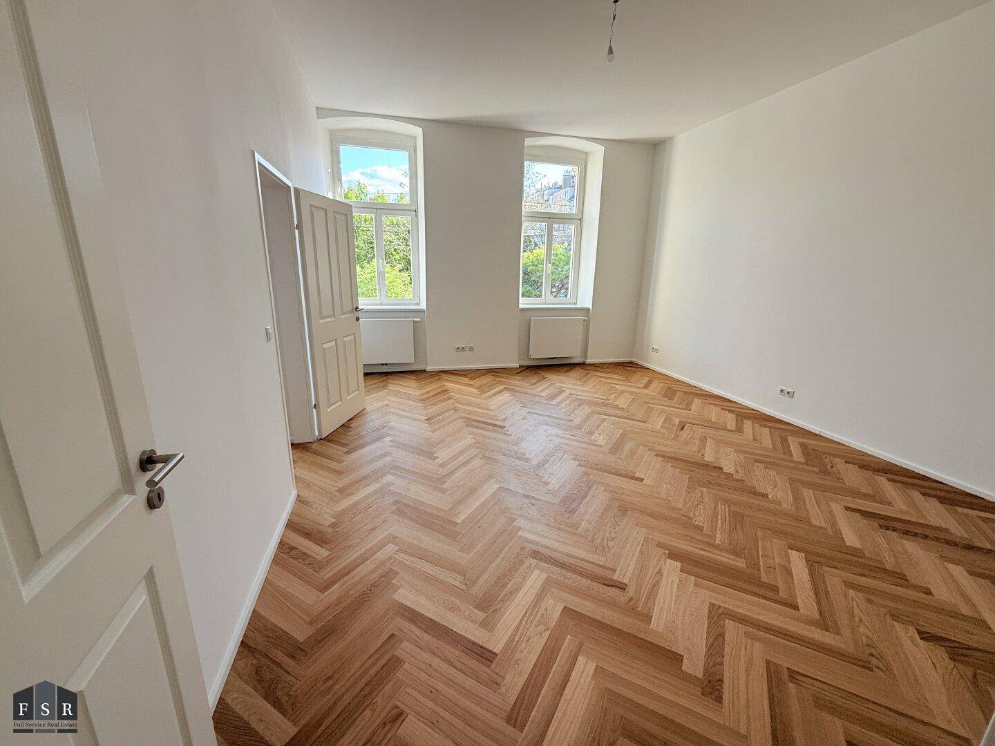 Großes, leeres Zimmer mit elegantem Fischgrätparkett und zwei hohen Fenstern.