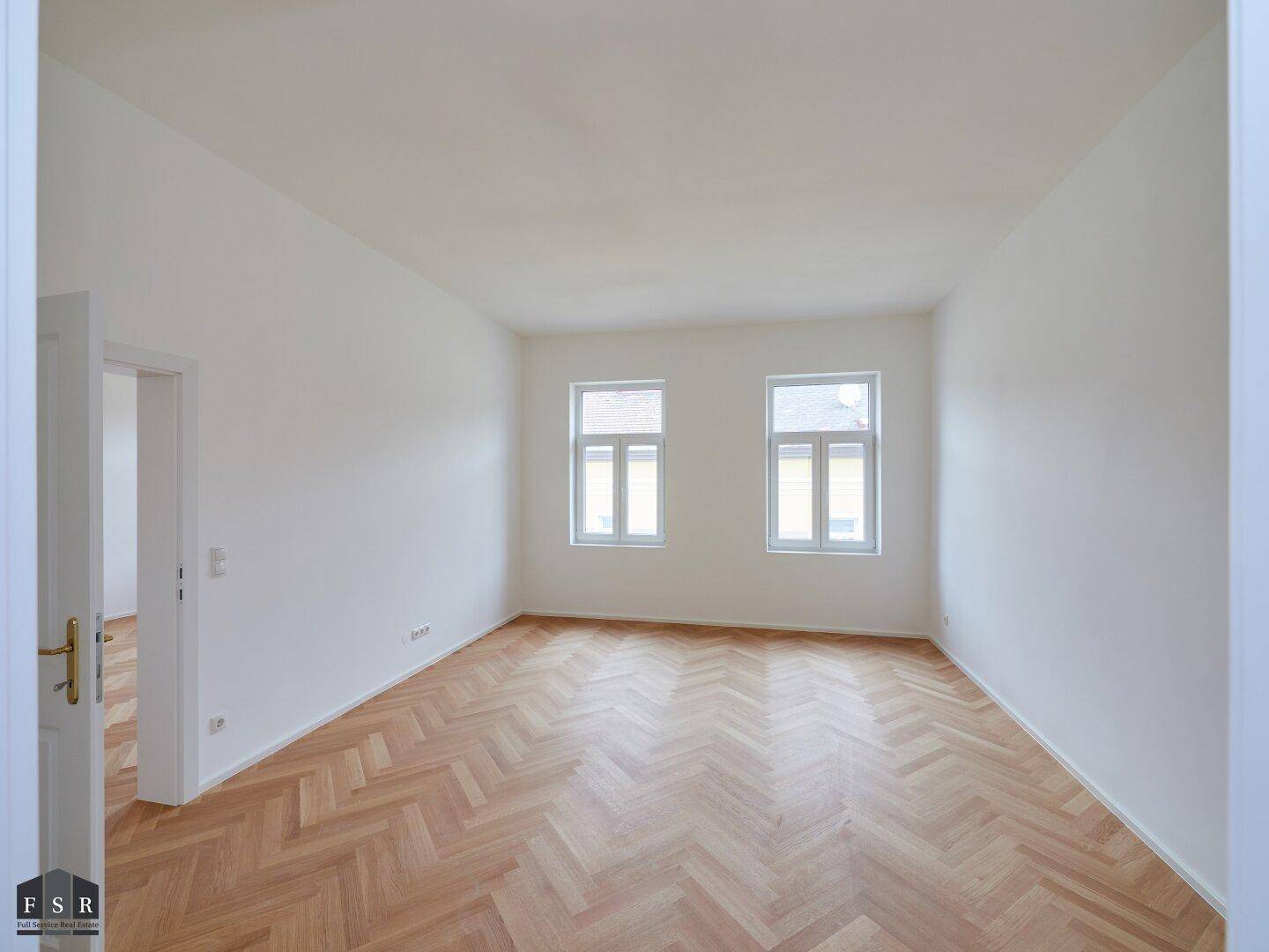 Geräumiges, helles Zimmer mit Fischgrätparkett und einer offenen Tür zu einem weiteren Raum.