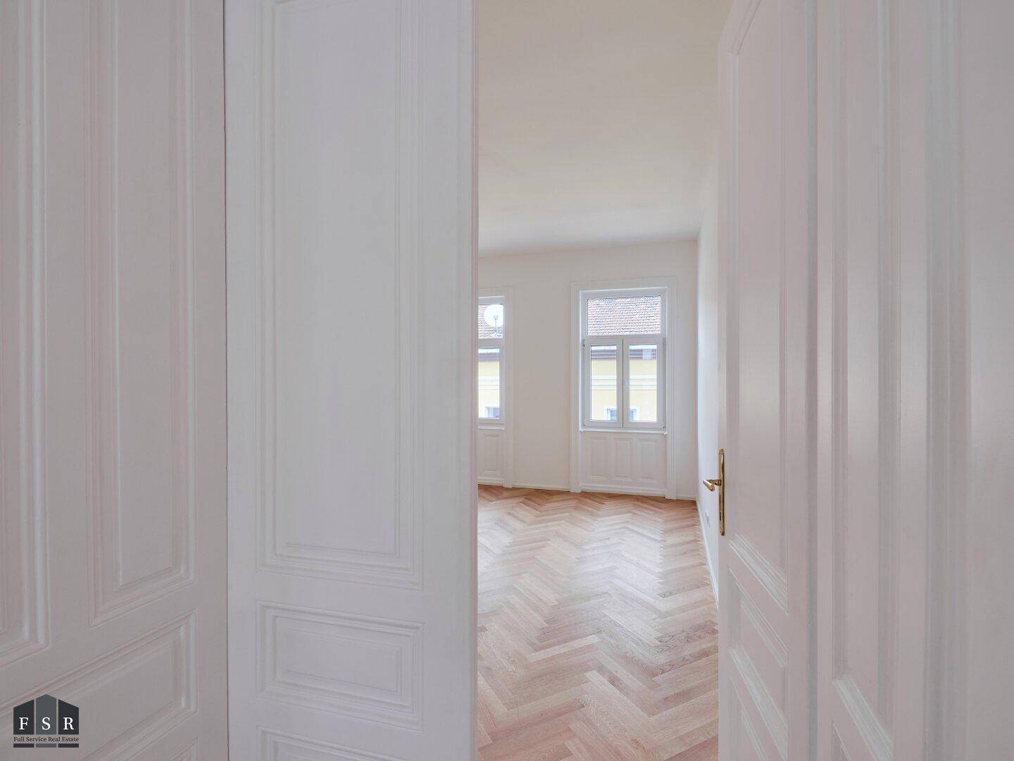 Blick durch weit geöffnete weiße Doppeltüren in ein helles Zimmer mit Fischgrätparkett.