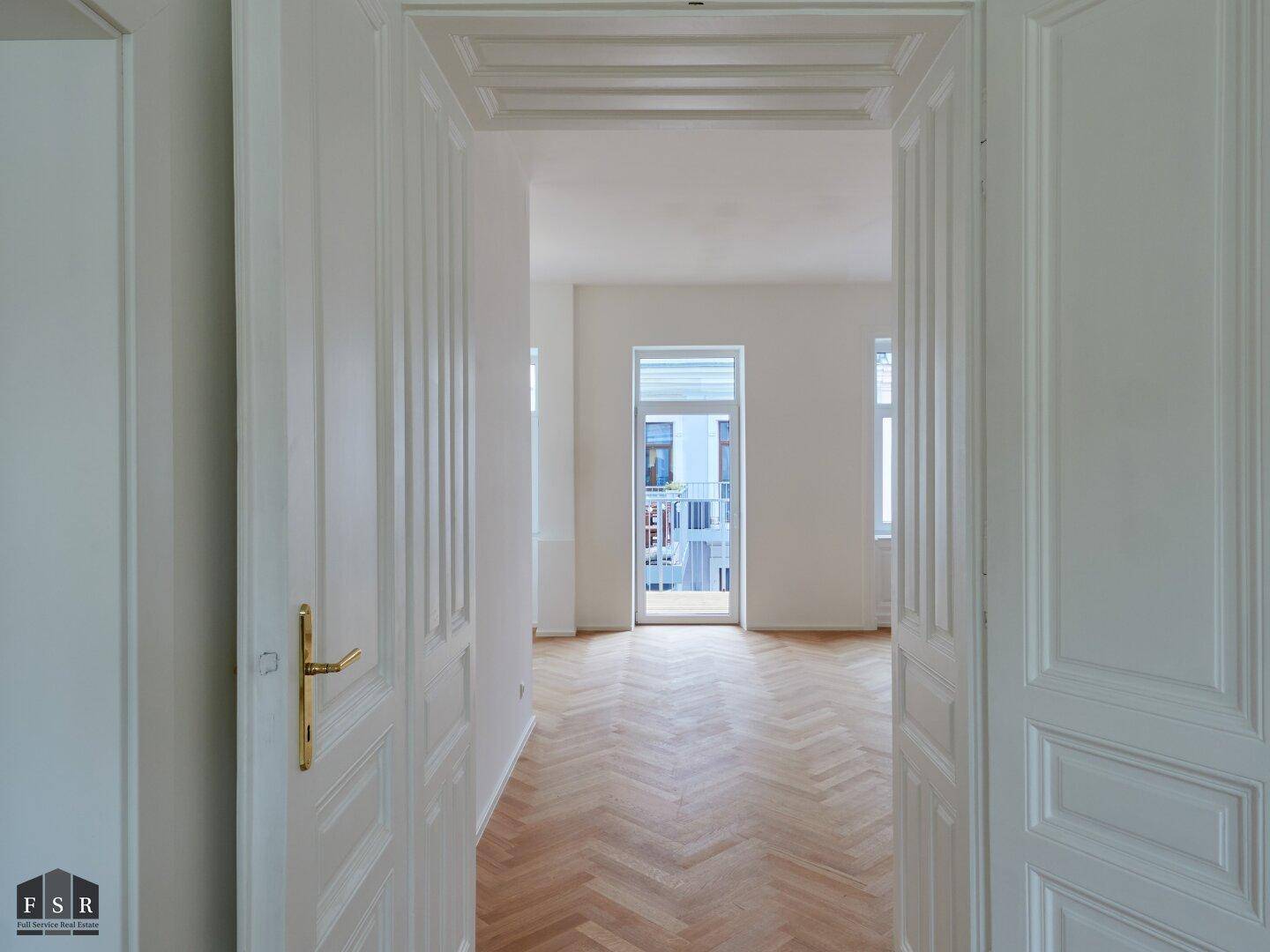 Blick durch eine Tür in einen hellen Raum mit Fischgrätparkett und Zugang zu einem Balkon.