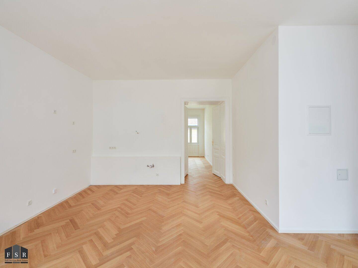 Leerer, heller Raum mit Fischgrätparkett und einer Tür, die in einen weiteren Bereich führt.