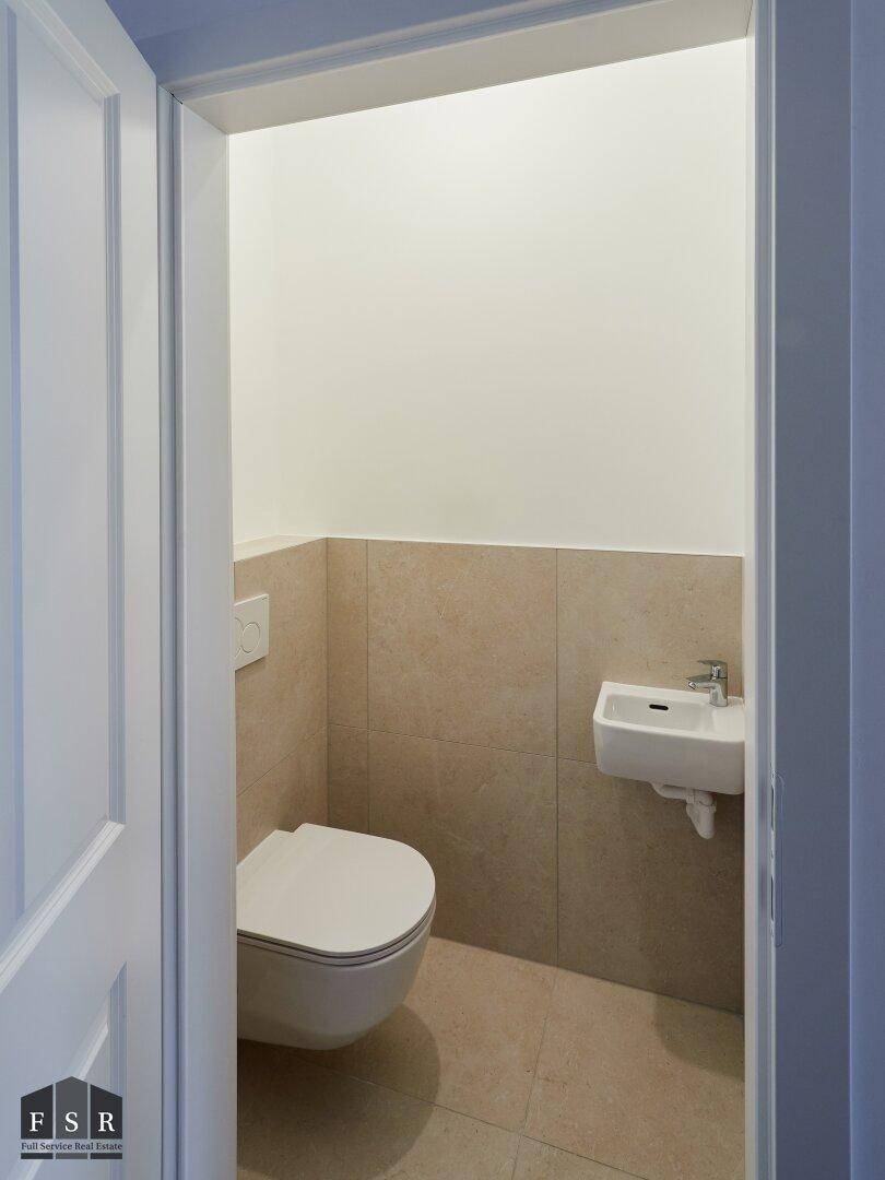 Kleines Gäste-WC mit wandhängender Toilette, kleinem Waschbecken und beigefarbenen Fliesen.
