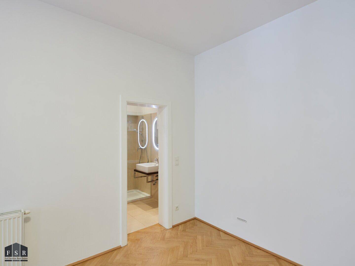 Helles Zimmer mit Parkettboden und Blick auf ein modernes Badezimmer durch die offene Tür.