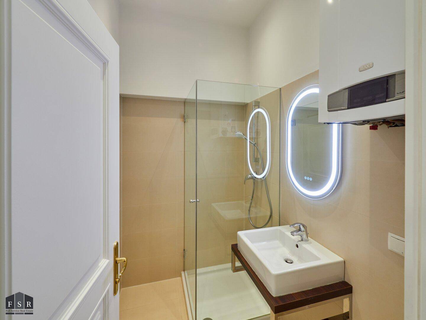 Elegantes Badezimmer mit Glasdusche, Waschtisch und beleuchtetem Spiegel, in hellen Tönen gehalten.