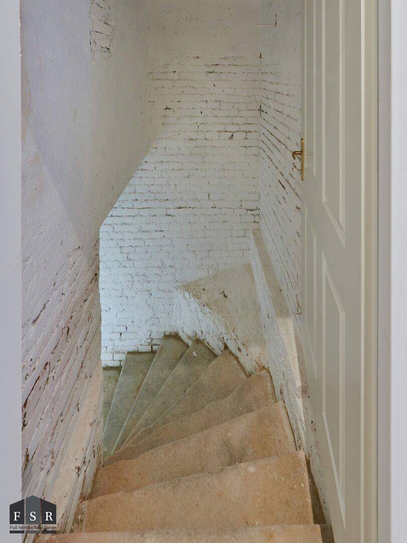 Steile Kellertreppe mit unverputzten Ziegelwänden, die in einen Lagerbereich führt.