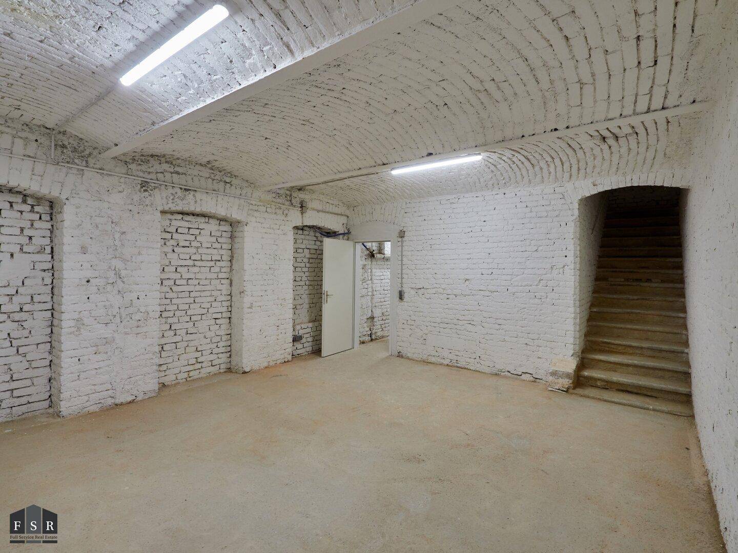 Großer Kellerraum mit Ziegelwänden, gewölbter Decke und Zugang zu weiteren Bereichen.