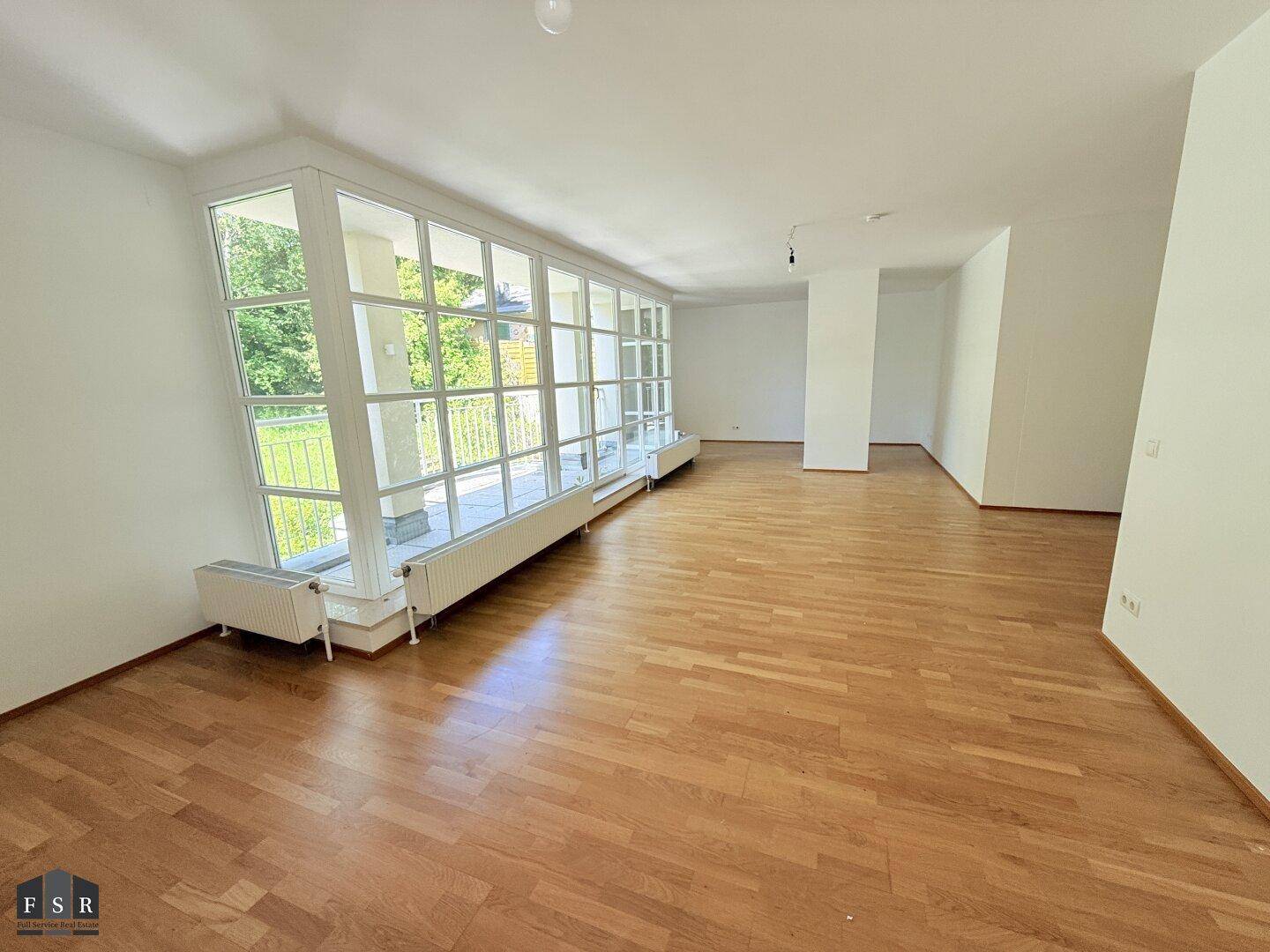 Weitläufiges Wohnzimmer mit Parkettboden und großen Fenstern, die viel Licht hereinlassen.