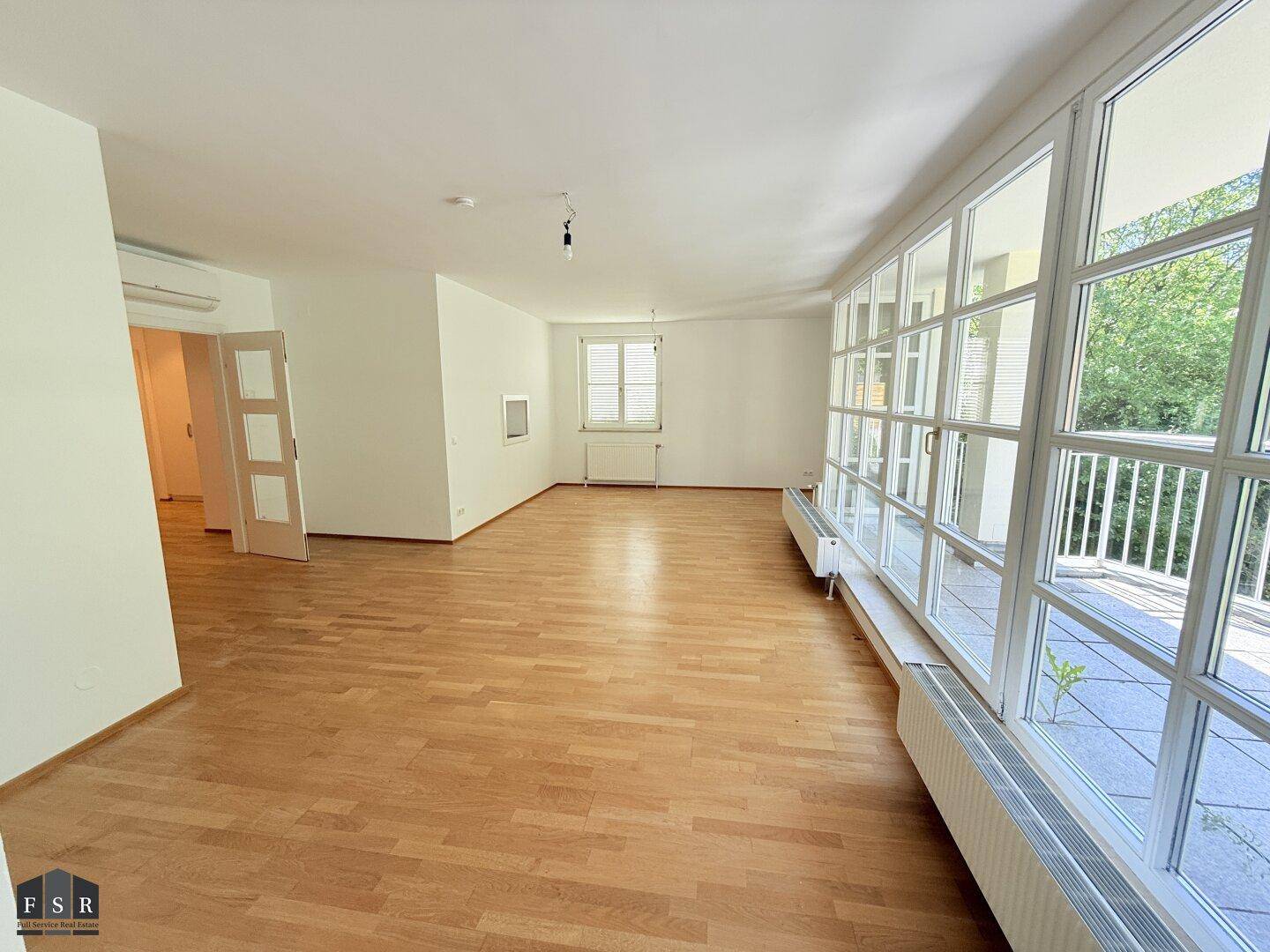 Geräumiges Wohnzimmer mit Holzboden, großen Fenstern und Zugang zu einem Balkon.
