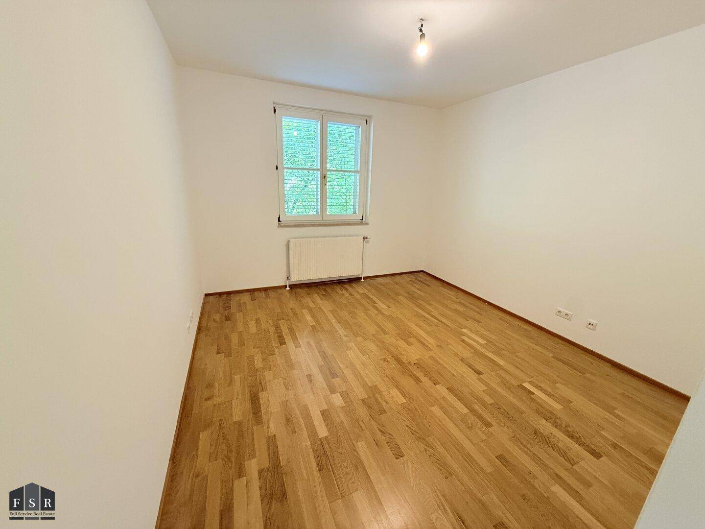 Helles Schlafzimmer mit Parkettboden, weißen Wänden und einem Fenster mit Jalousien.