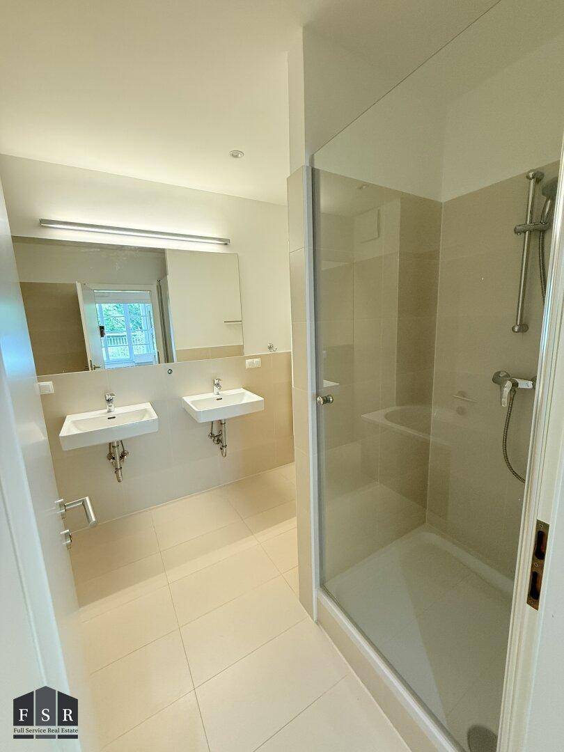 Modernes Badezimmer mit Doppelwaschbecken, Dusche und hellen Fliesen an Wänden und Boden.
