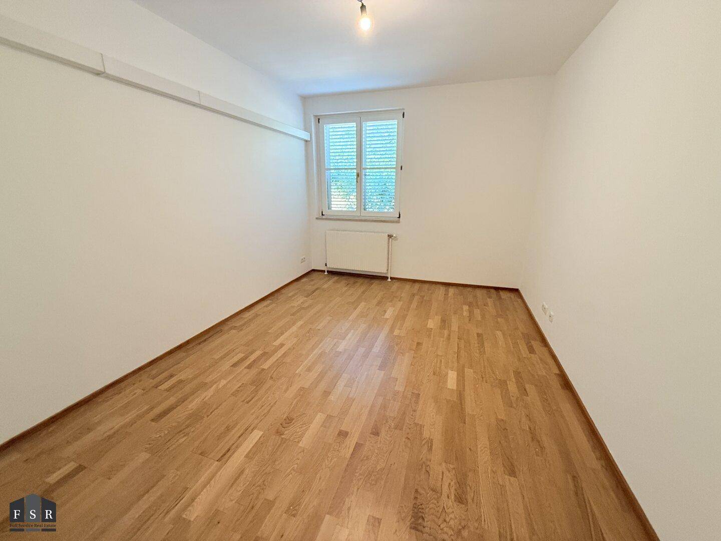 Schlafzimmer mit Parkettboden, weißen Wänden und einem Fenster mit Jalousien.
