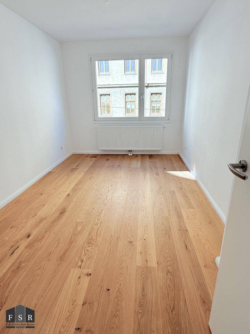 Unmöbliertes Zimmer mit hellem Parkettboden und Blick auf die gegenüberliegenden Gebäude.