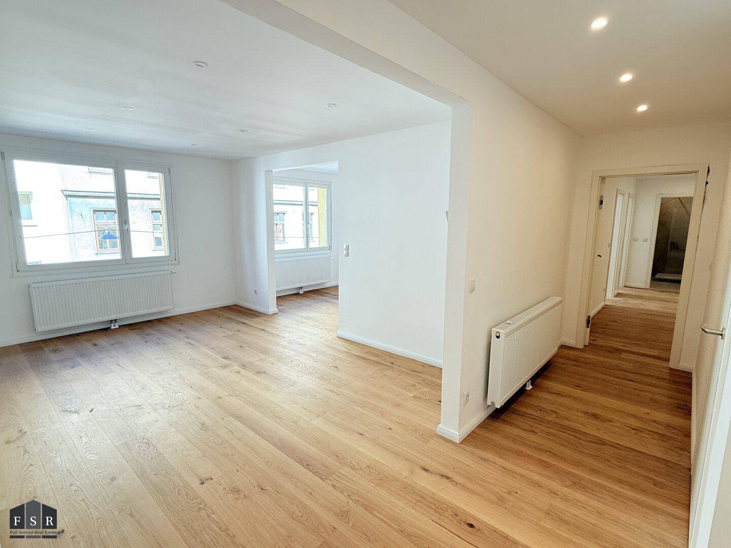 Geräumiges Wohnzimmer mit hellem Parkettboden, weißen Wänden und viel Tageslicht.