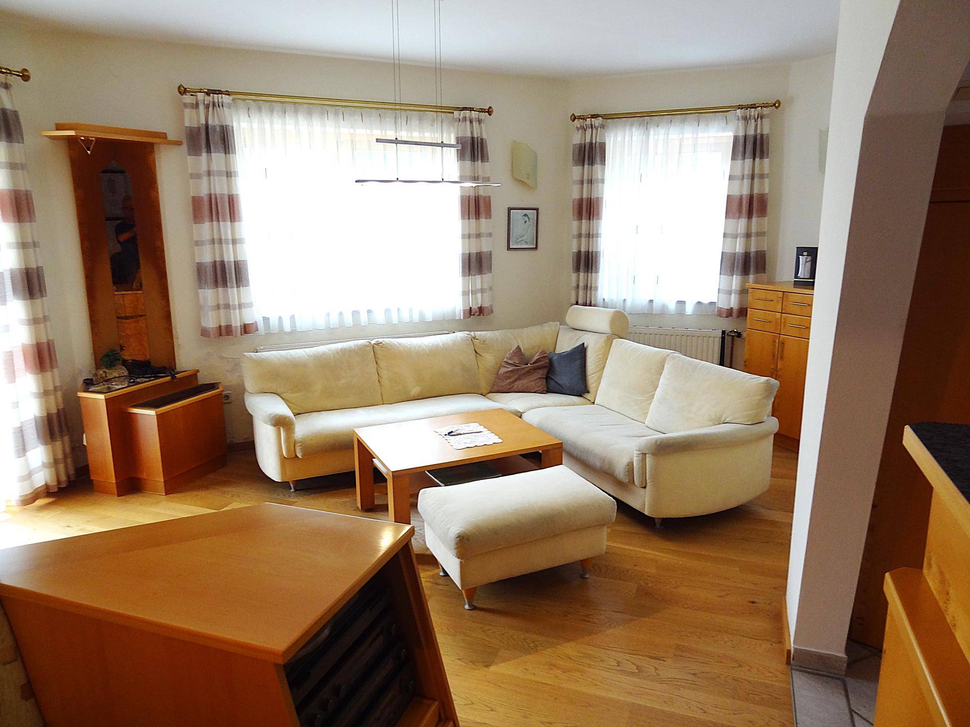 Gemütliches Wohnzimmer mit hellem Ecksofa, Couchtisch und großen Fenstern.