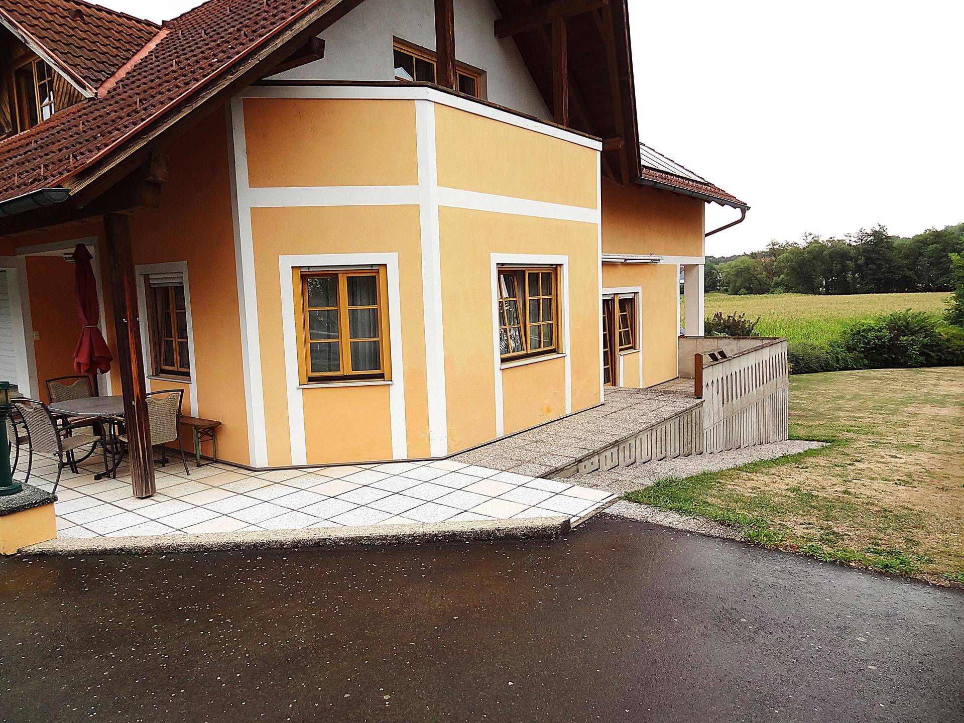 Hausfassade in Gelb mit Terrasse, Rampe und Blick auf grüne Felder.