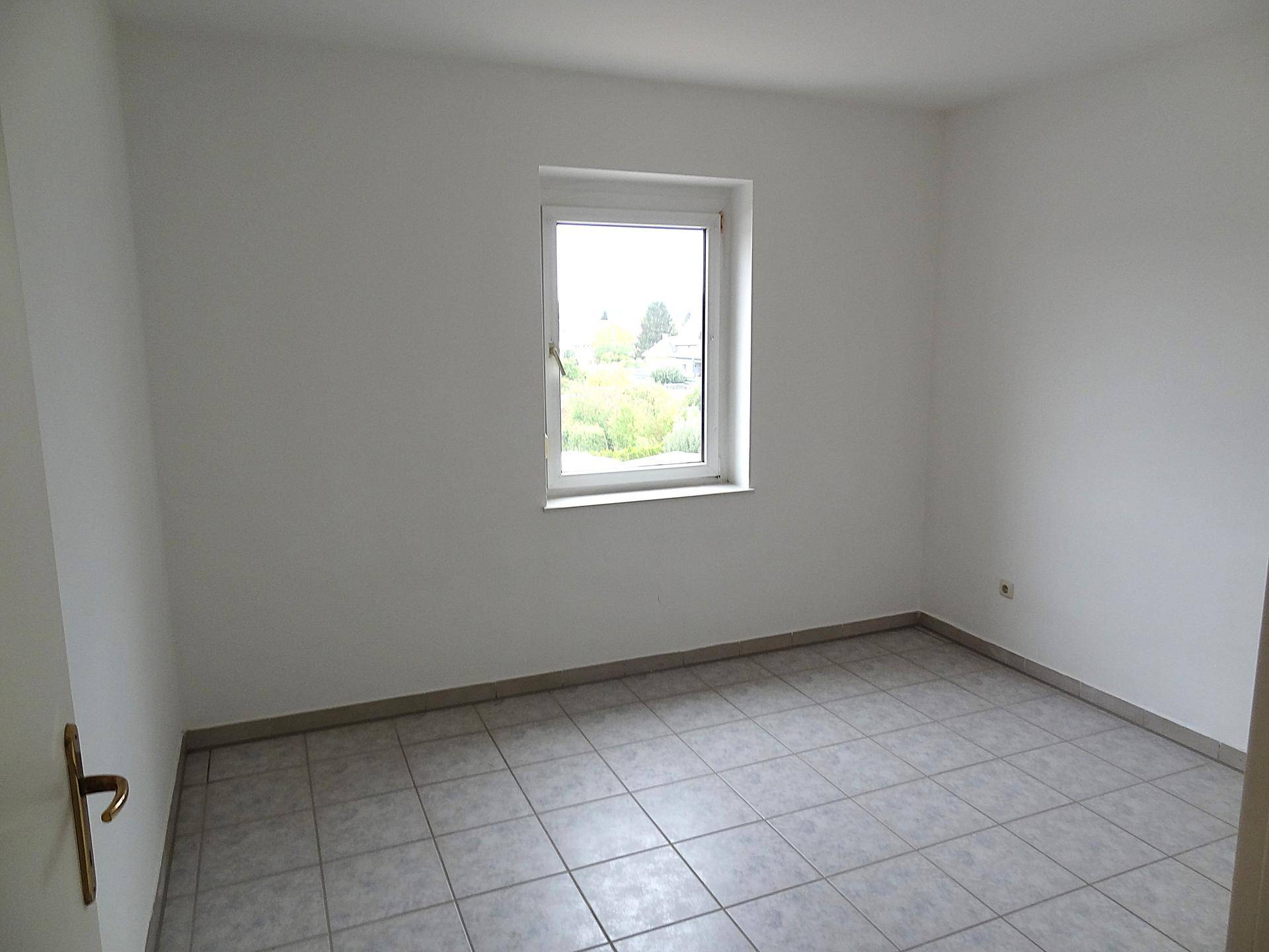 Unmöbliertes Zimmer mit Fliesenboden, weißen Wänden und einem Fenster mit Ausblick ins Grüne.