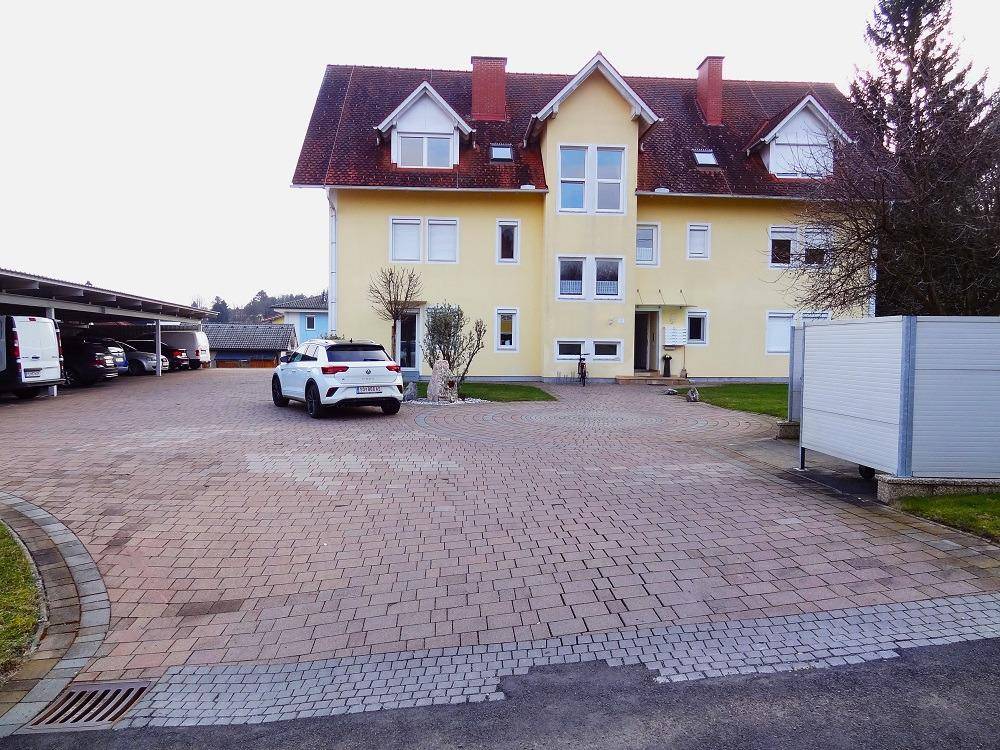 Gelbes Mehrfamilienhaus mit rotem Dach und großem gepflasterten Parkplatzbereich im Freien.