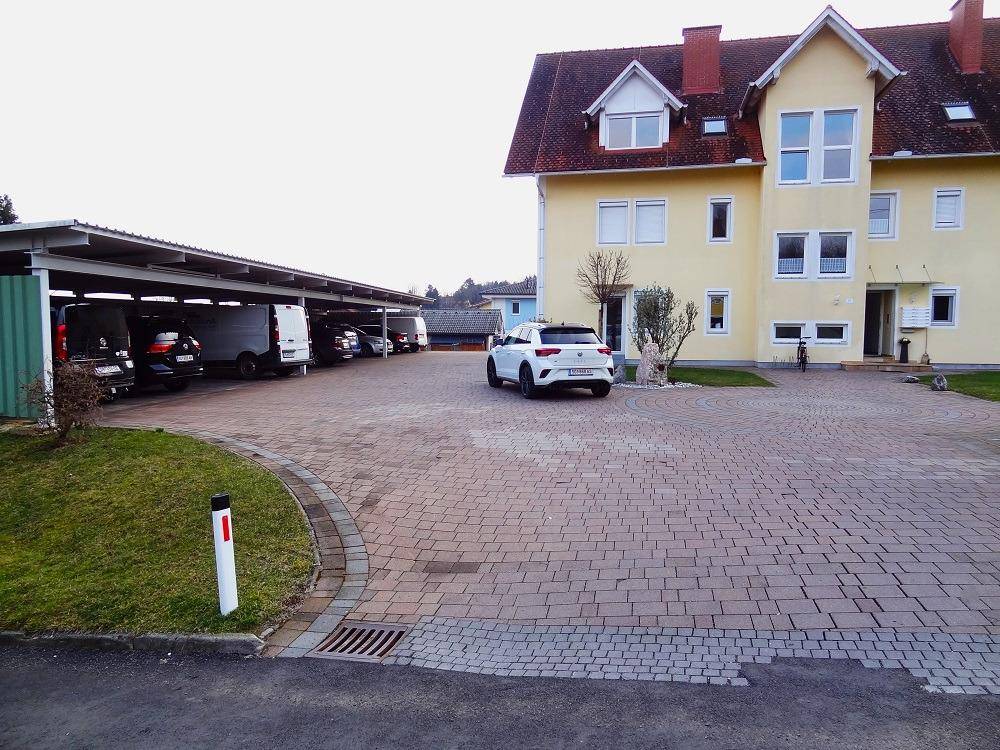 Außenansicht des Gebäudes mit großem gepflasterten Parkplatz und einem überdachten Carportbereich.