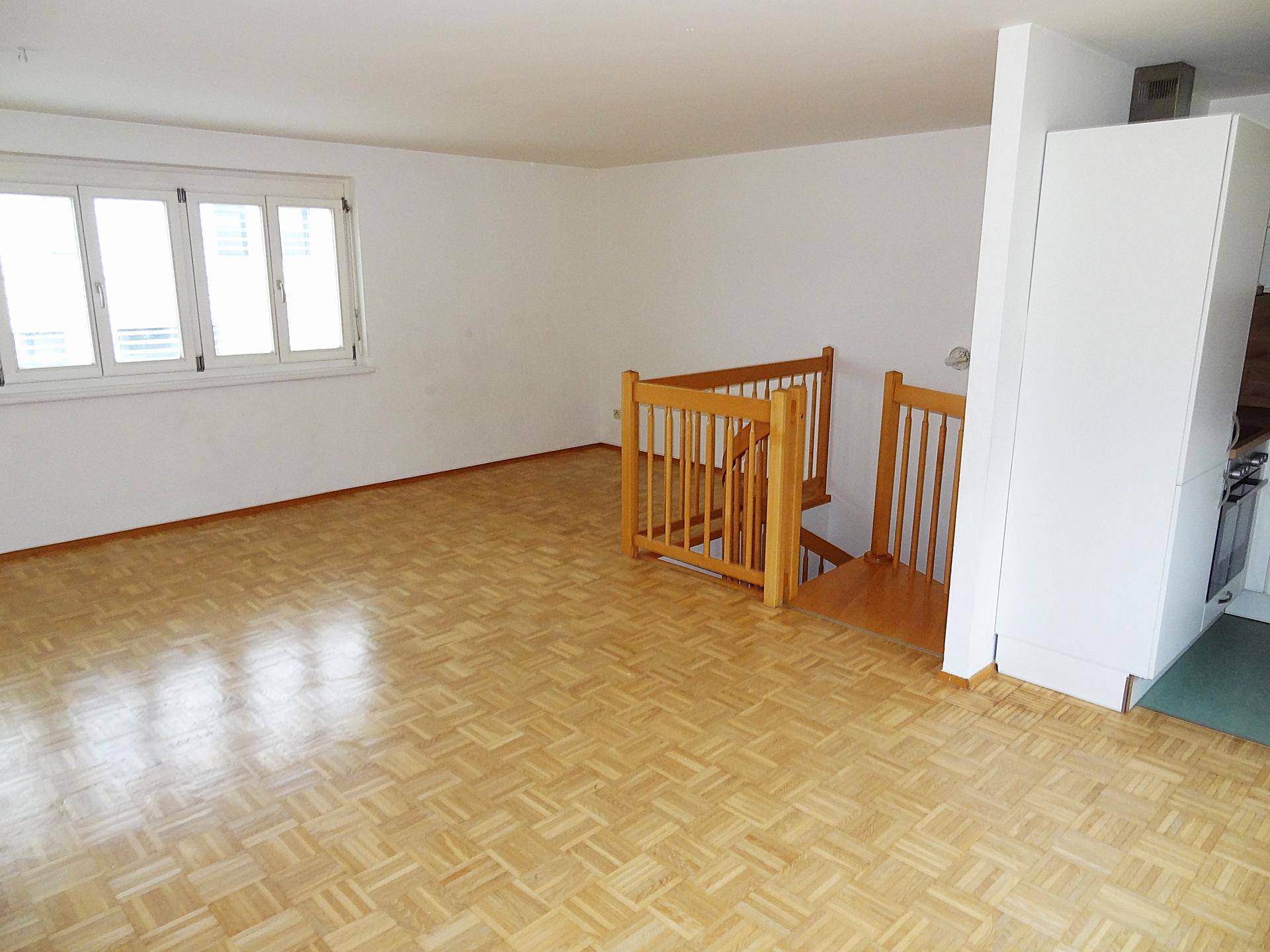 Geräumiges Wohnzimmer mit hellem Parkettboden, großen Fenstern und Holztreppe.