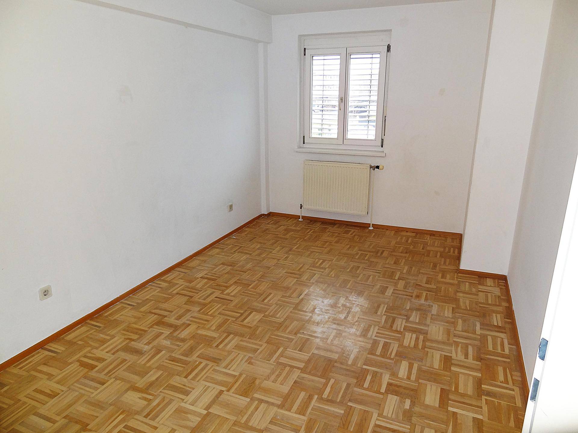 Helles Schlafzimmer mit Parkettboden, weißen Wänden und Fenster mit Heizkörper.