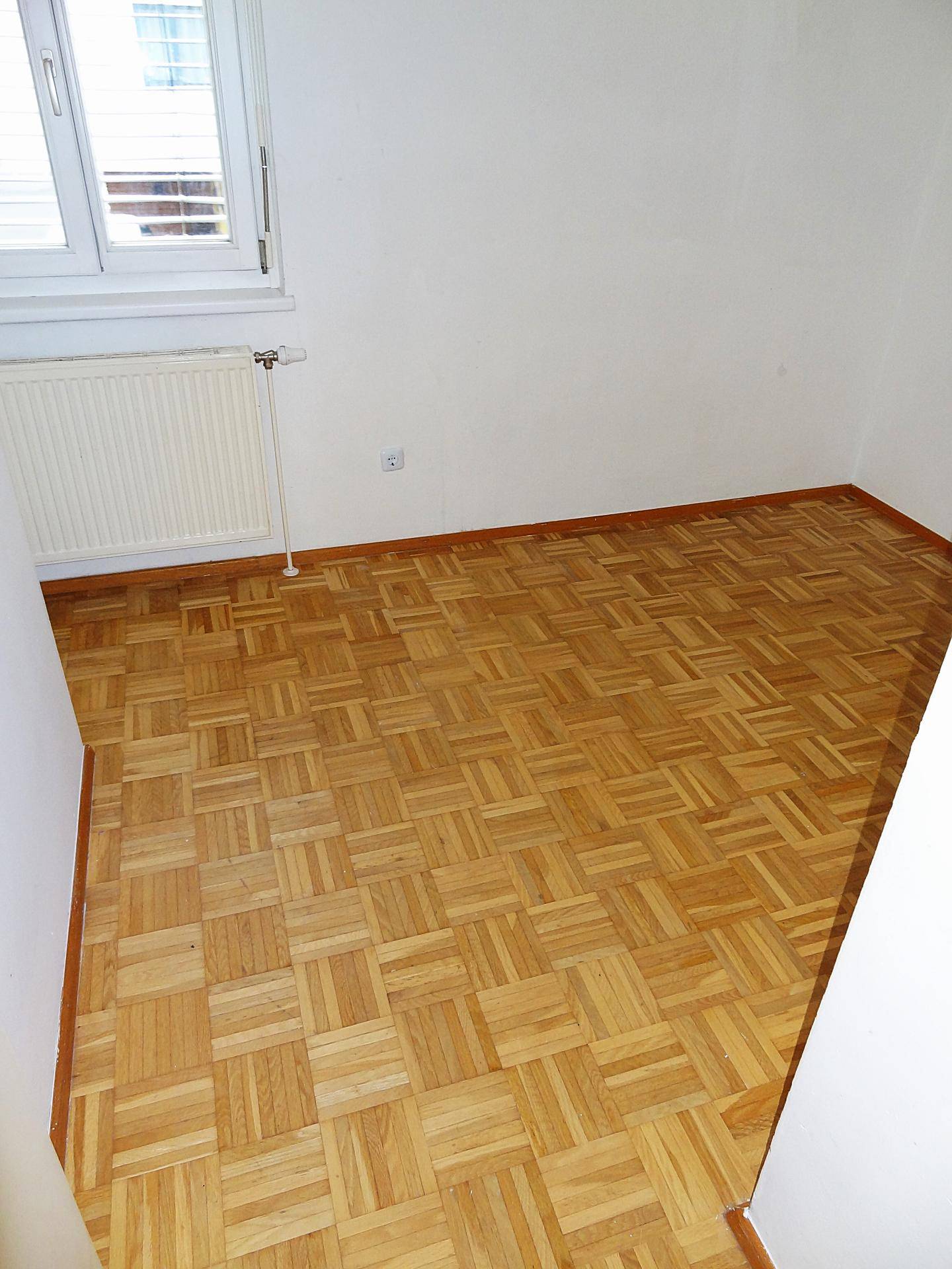 Leerer Raum mit hellem Parkettboden, weißen Wänden und einem Fenster mit Heizkörper.