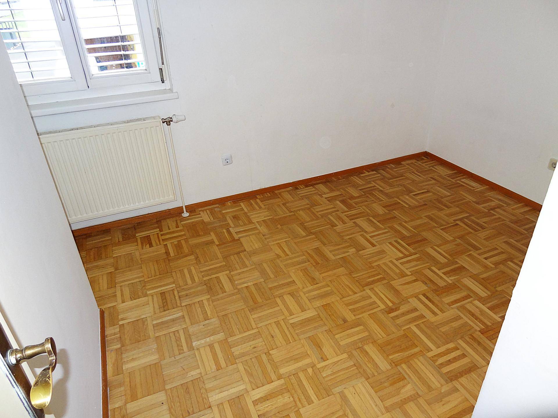 Heller Raum mit Parkettboden, weißen Wänden und einem Fenster mit Heizkörper.