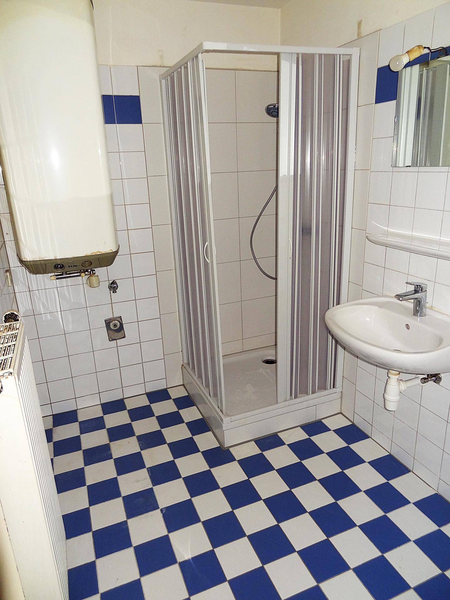 Badezimmer mit Duschkabine, Waschbecken und Warmwasserboiler, auf blau-weißem Fliesenboden.