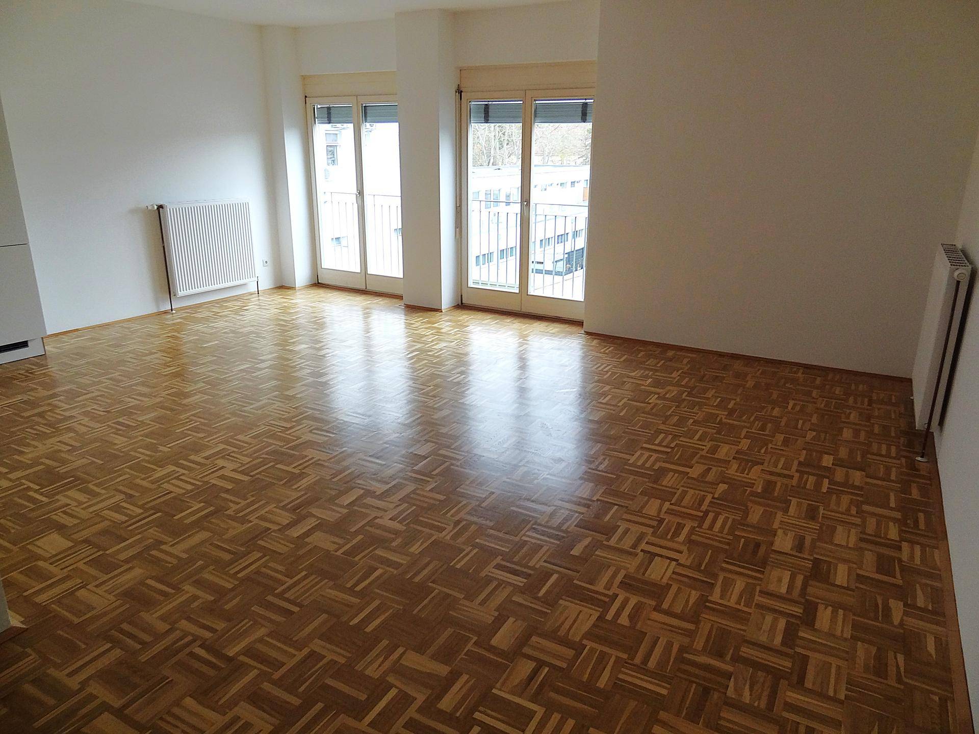 Geräumiger Wohnraum mit hellem Parkettboden und Zugang zu einem Balkon durch Fenstertüren.