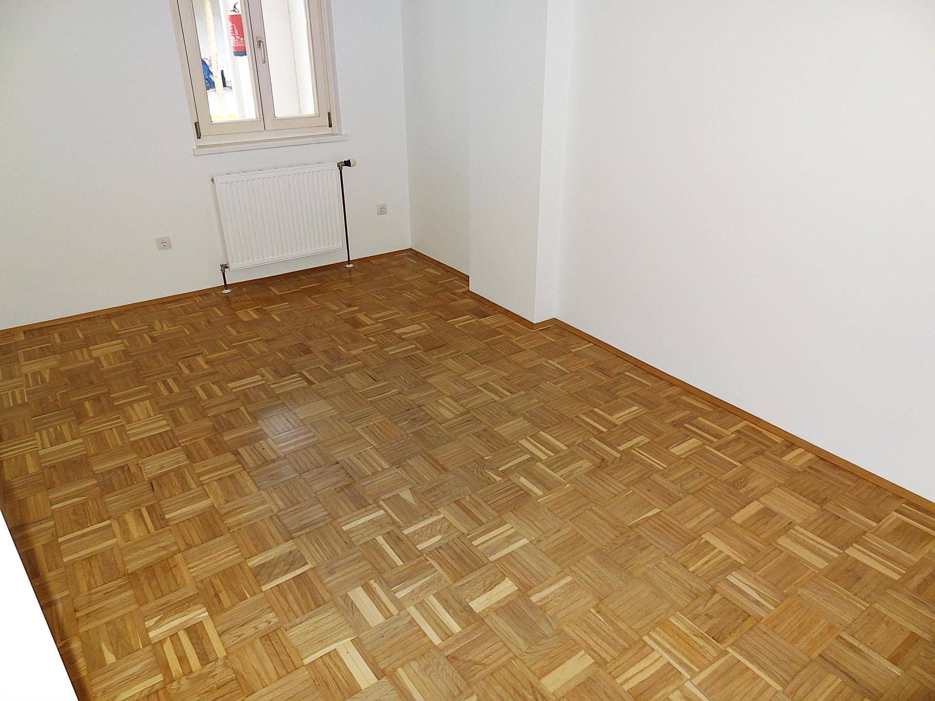 Leeres Zimmer mit hellem Parkettboden, weißen Wänden und einem Fenster mit Heizkörper.