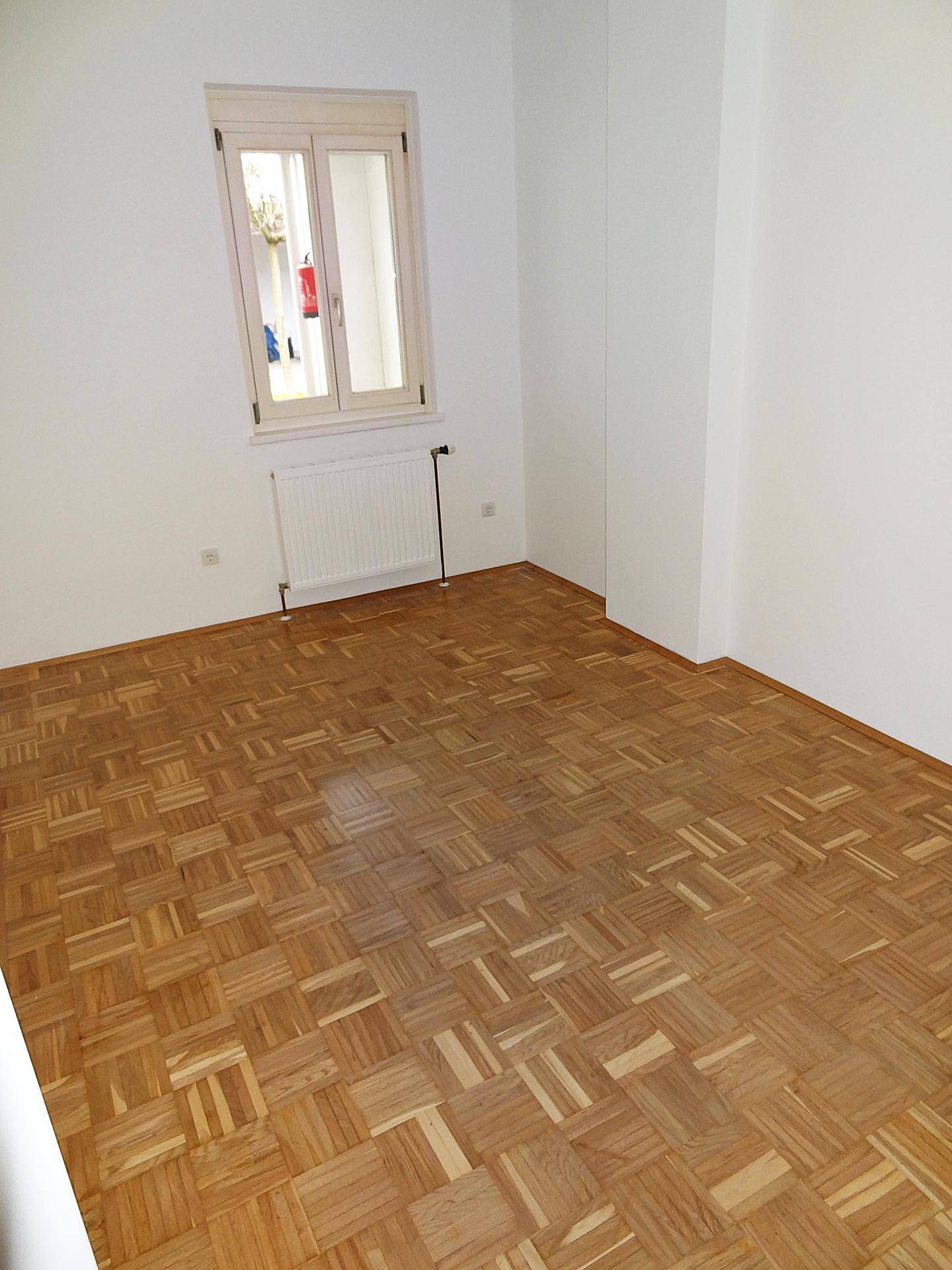 Helles Schlafzimmer mit Parkettboden, weißen Wänden und einem Fenster, das viel Licht hereinlässt.