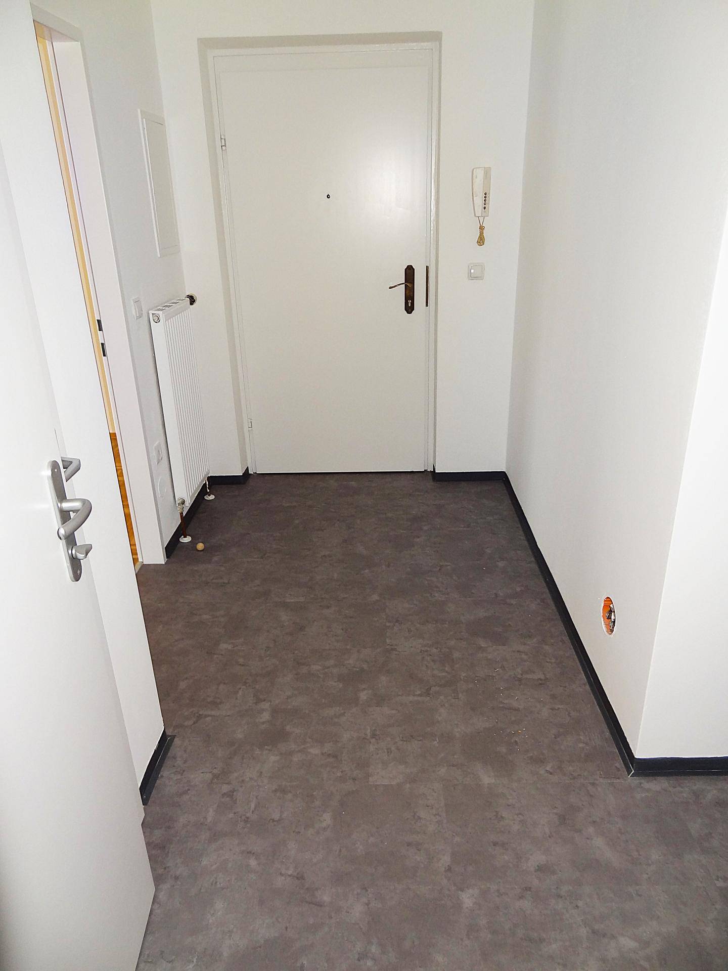 Eingangsbereich mit grauem Bodenbelag, weißer Wand, Gegensprechanlage und einem Heizkörper.