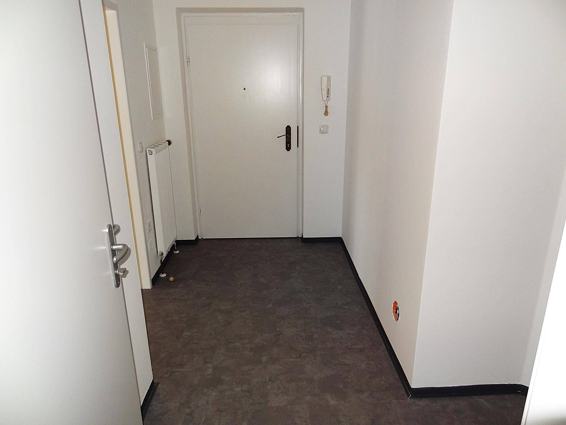 Eingangsbereich mit grauem Bodenbelag, weißer Wand, Gegensprechanlage und einem Heizkörper.