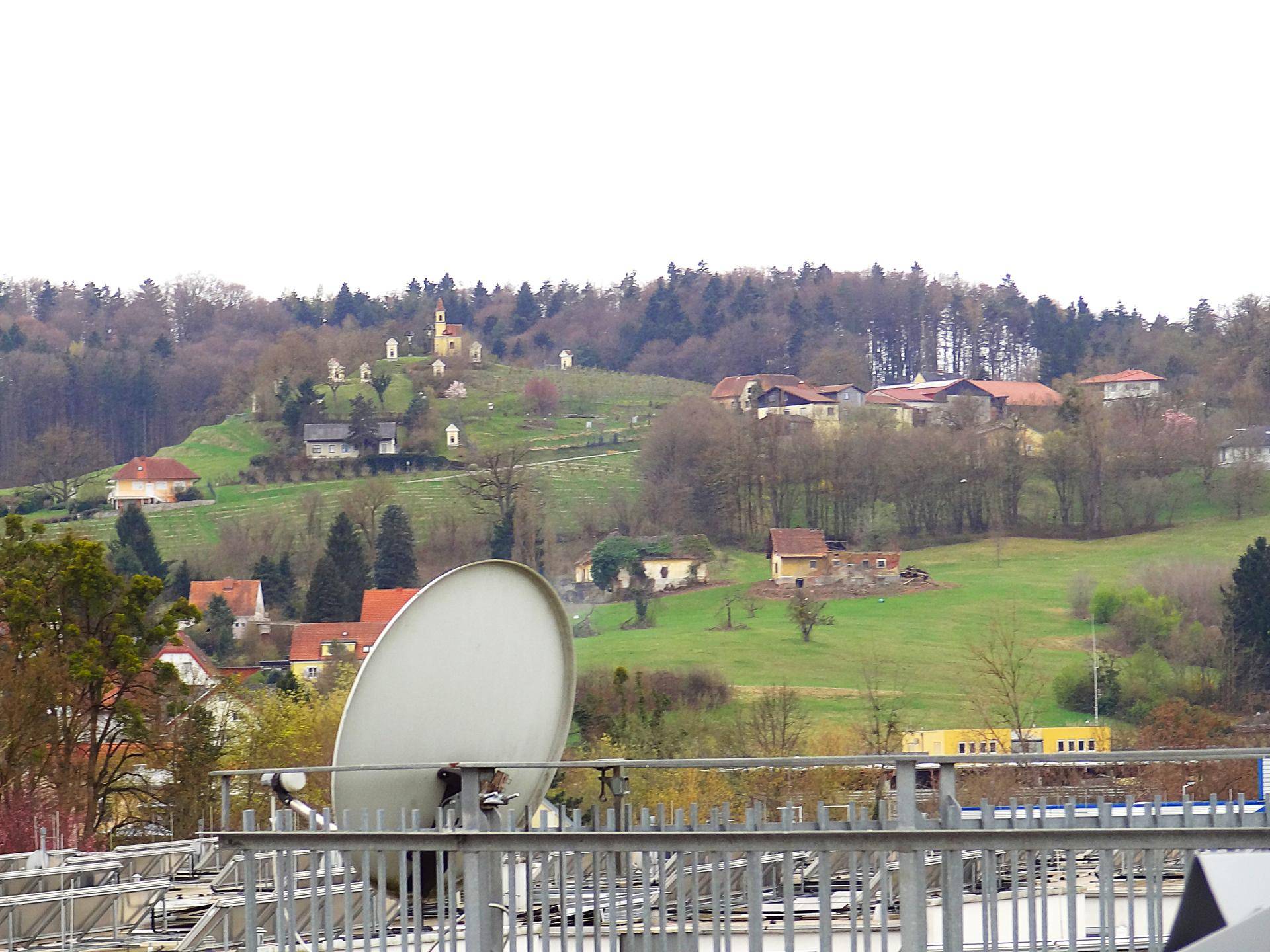 Weitblick über eine hügelige Landschaft mit Häusern und Bäumen, im Vordergrund eine Satellitenschüssel.