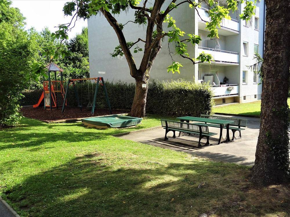 Gepflegter Gemeinschaftsgarten mit Spielplatz, Sitzgelegenheiten und Blick auf das Mehrfamilienhaus mit Balkonen.