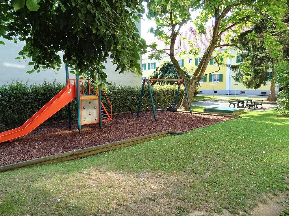 Grüner Gemeinschaftsgarten mit Kinderspielplatz, Rutsche, Schaukeln und Sandkasten vor einem Wohngebäude.