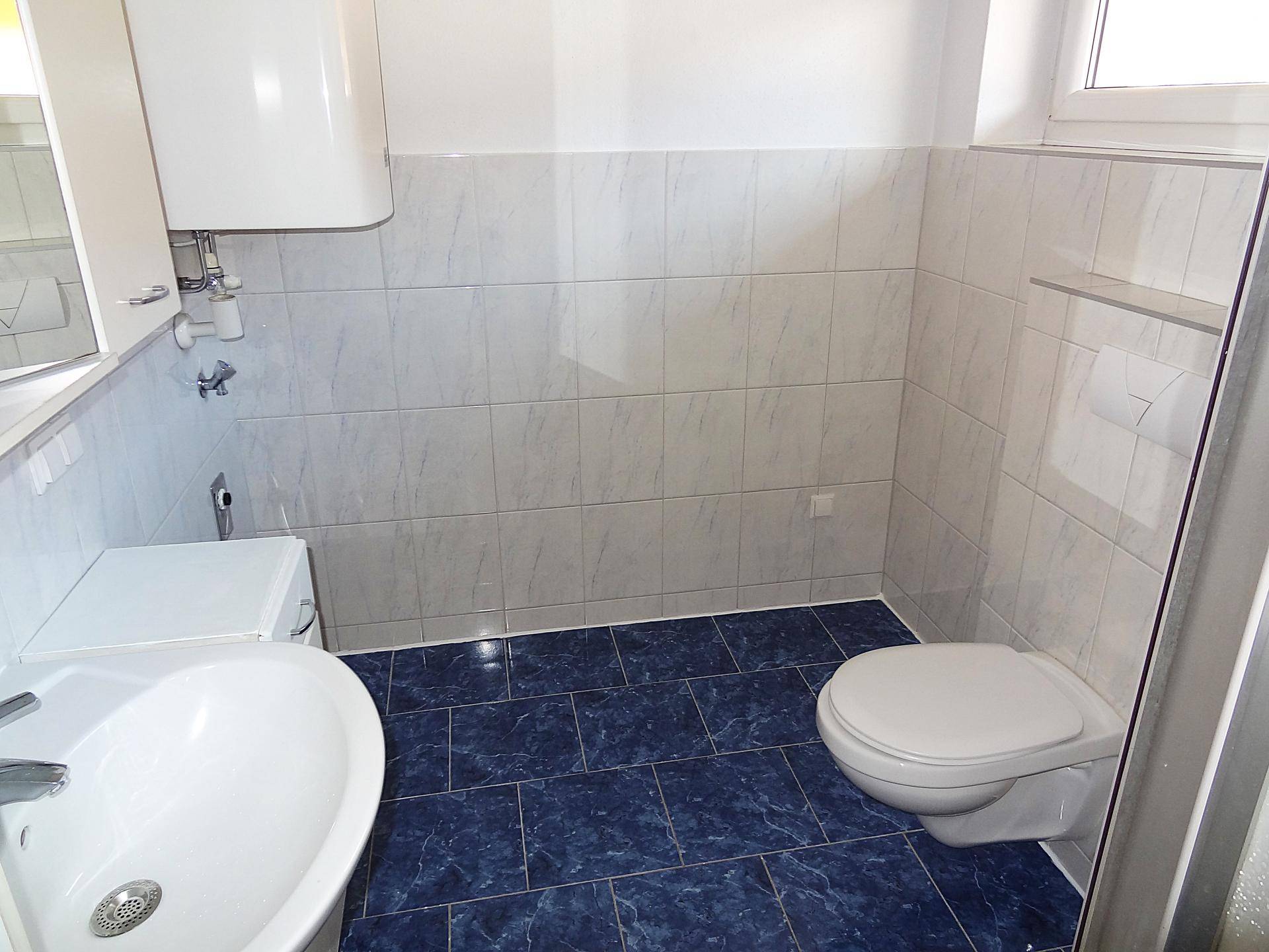 Badezimmer mit blauen Bodenfliesen, wandhängendem WC, Waschtisch und Fenster für Tageslicht.