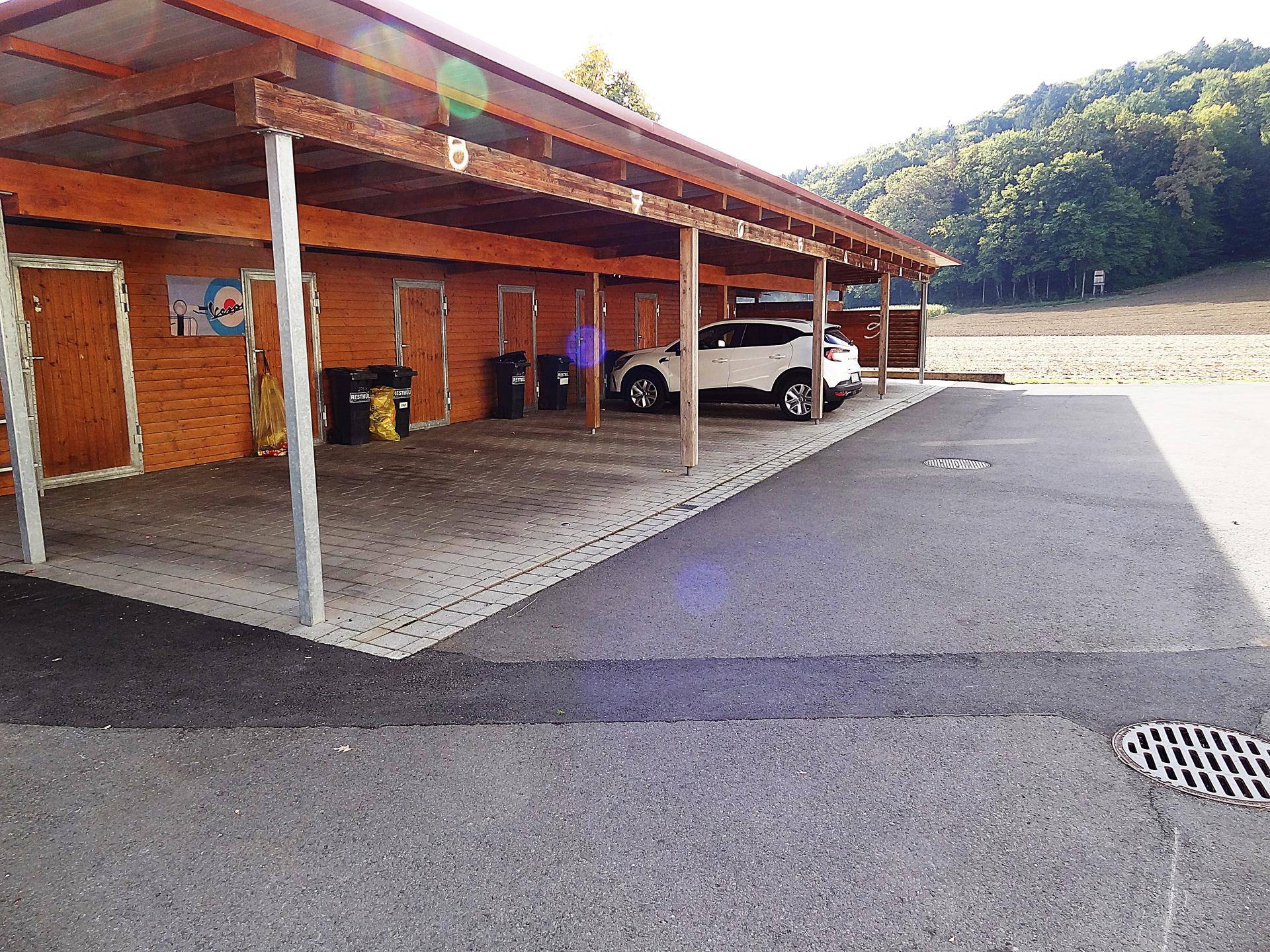 Überdachter Carport-Bereich mit Holzkonstruktion und einzelnen Abstellräumen, inklusive Parkplatz.