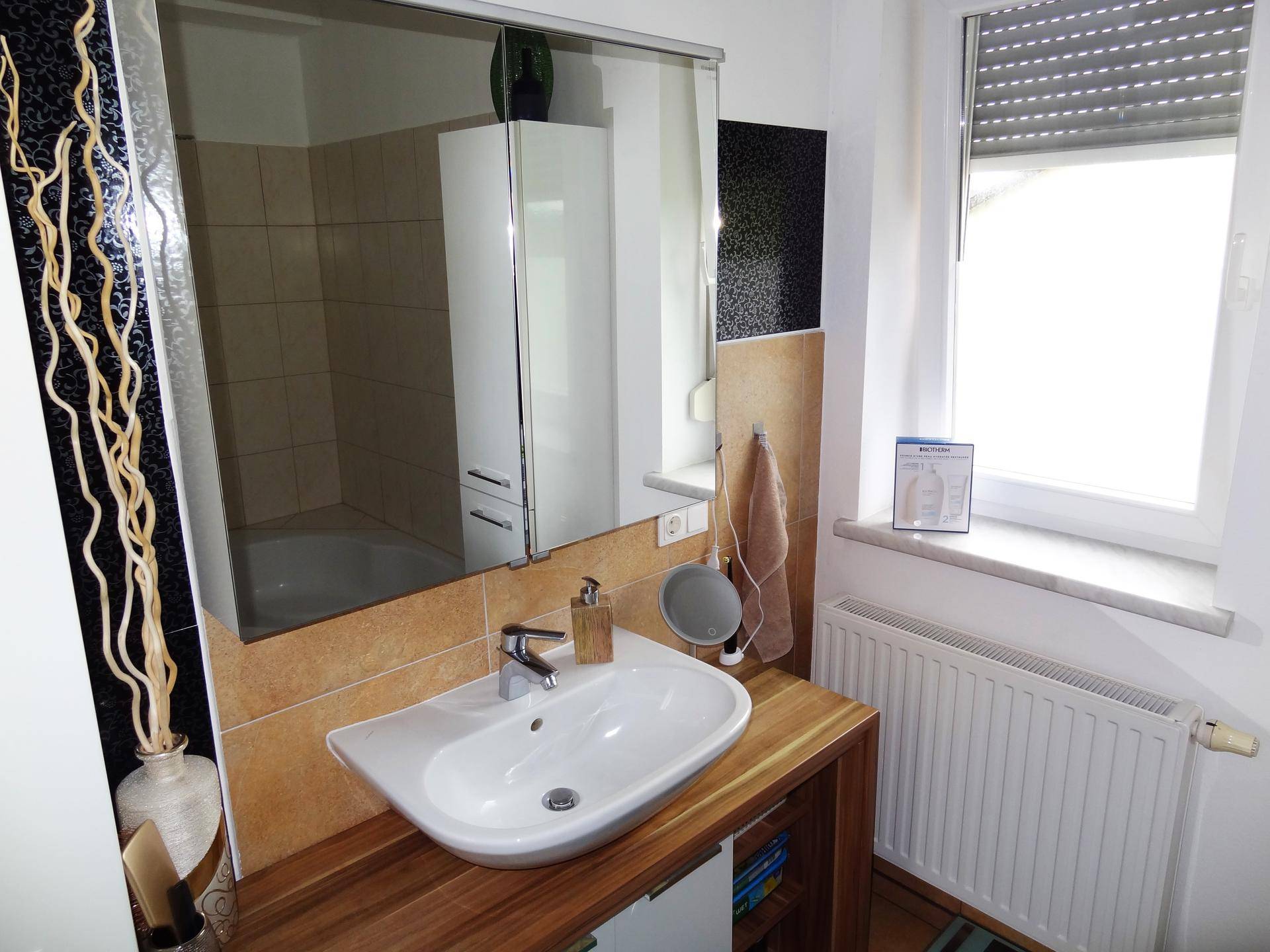 Waschbeckenbereich im Badezimmer mit Holzunterschrank, Spiegelschrank und Fenster für natürliche Helligkeit.