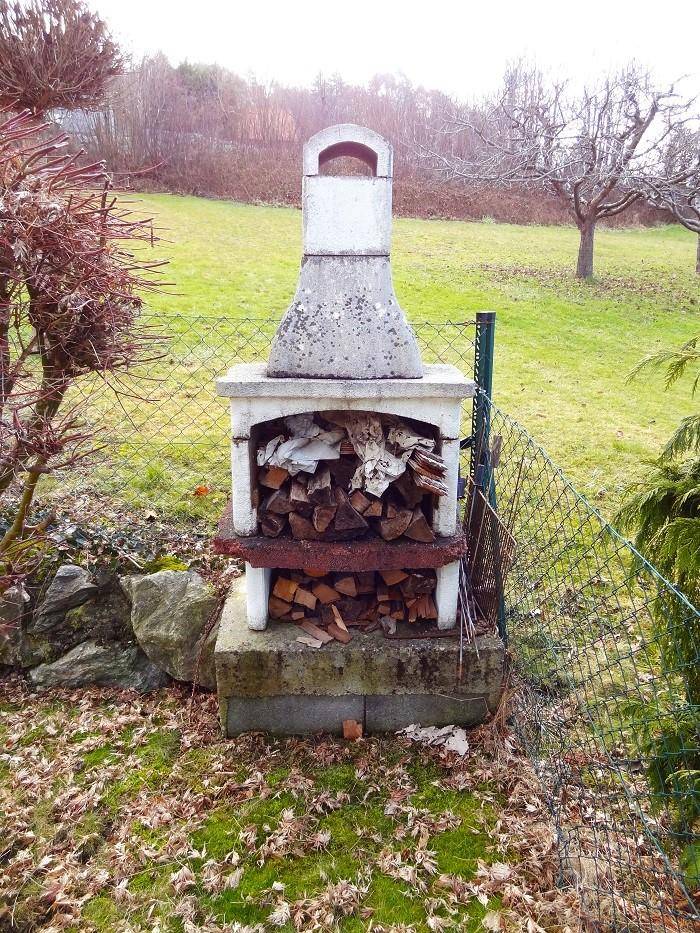 Rustikaler Außenkamin mit Holzscheiten und Grillbereich in einem grünen Garten.